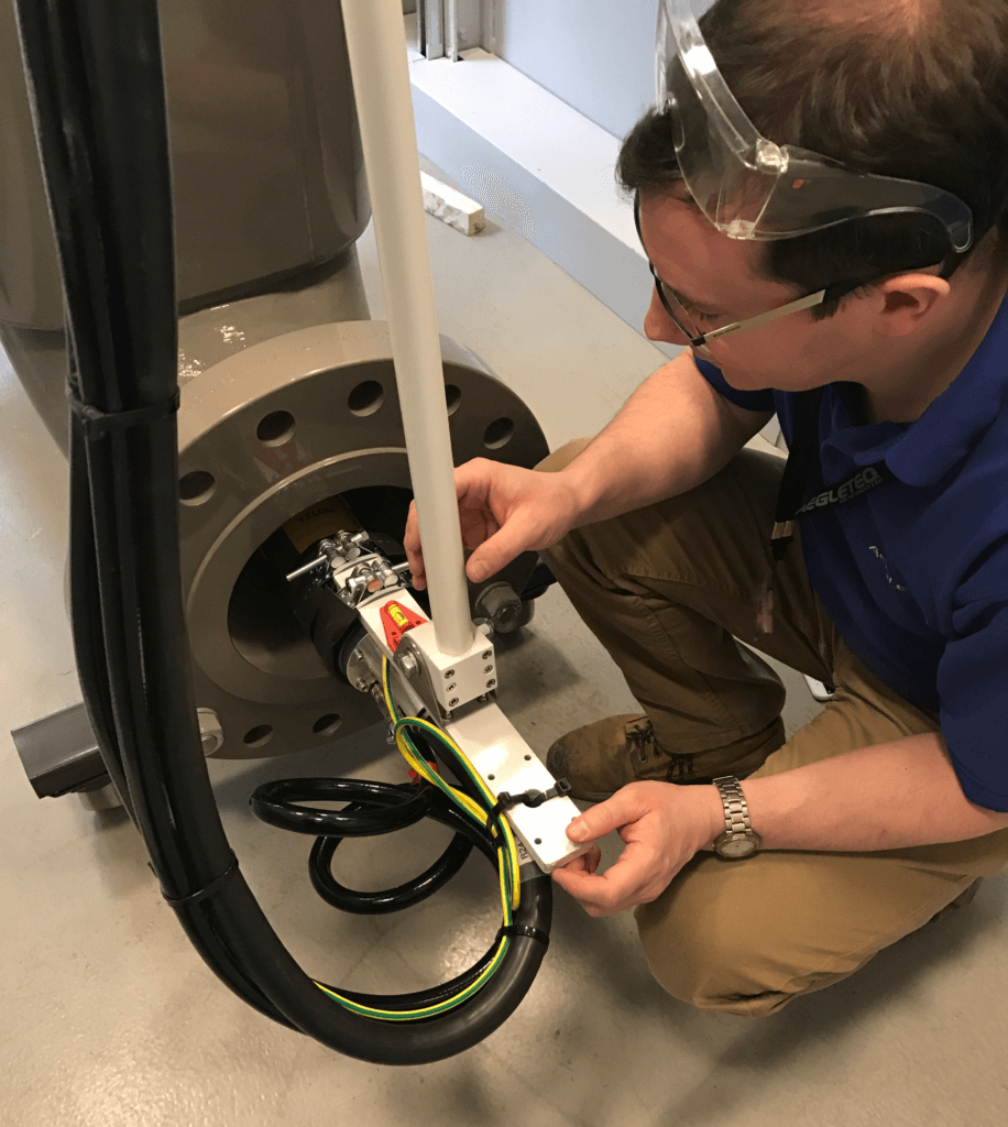 Een man knielt naast een apparaat met een grote buis. Hij werkt aan de onderzijde, met focus op draden en een mechanische schakelaar.