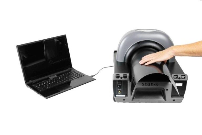 Een hand plaatst iets in een ronde opening van een röntgenapparaat, verbonden met een laptop. Het apparaat is ontworpen voor het scannen van objecten.